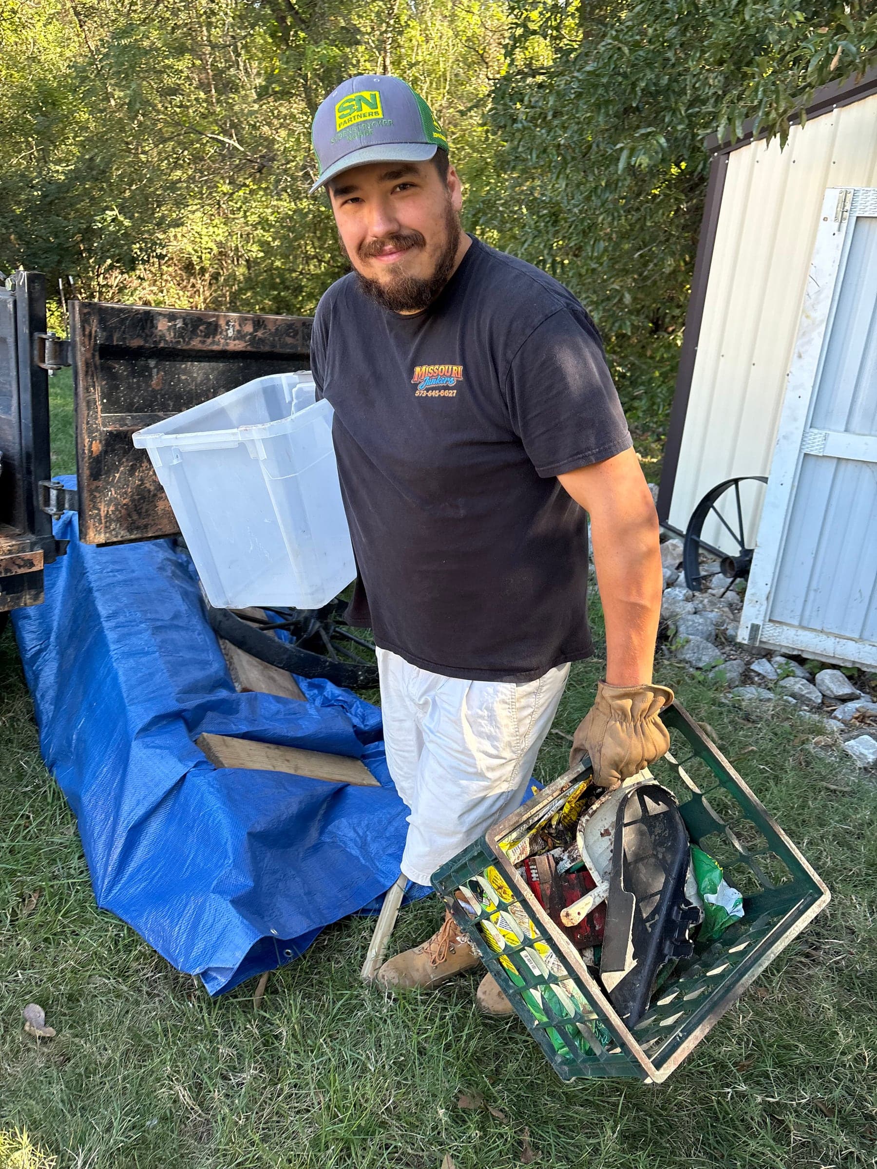 Cole carrying a crate of scrap to the dump trailer beside a metal shed