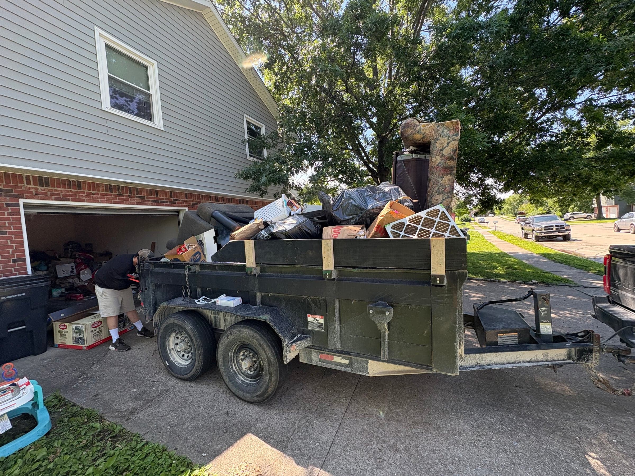 A dump trailer loaded with furniture and boxes in a driveway beside a brick house